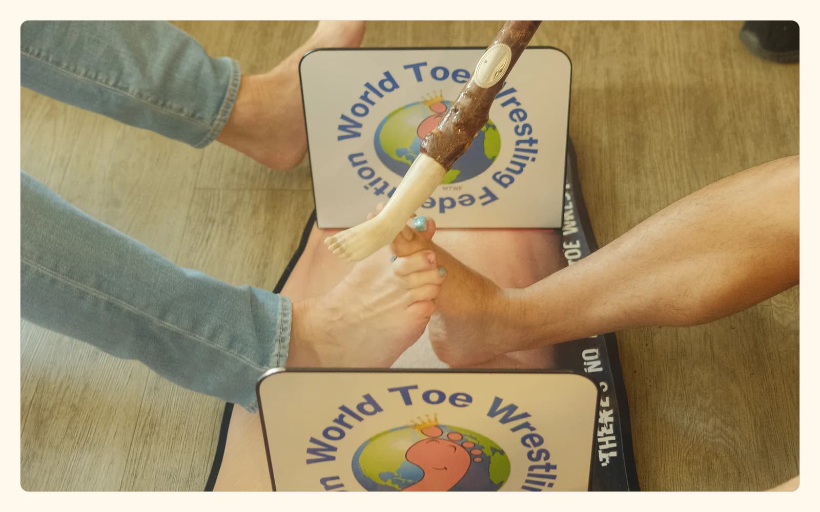 Two competitors interlock their feet inside a small toe-wrestling arena marked with World Toe Wrestling Federation signs.