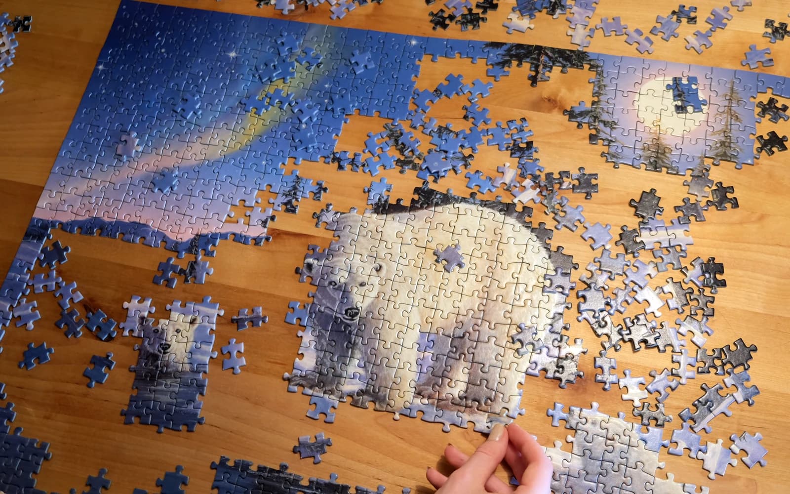 A partly assembled blue and cream jigsaw puzzle on a wooden table, surrounded by loose pieces and a hand placing one piece.