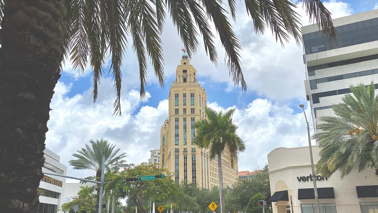 A palm-lined Coral Gables street with a tall yellow building under a bright, cloudy sky.