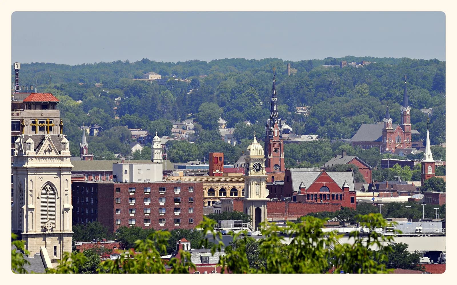 A view across downtown Dubuque, Iowa, with church spires, red brick buildings, a clock tower, and tree-covered bluffs.