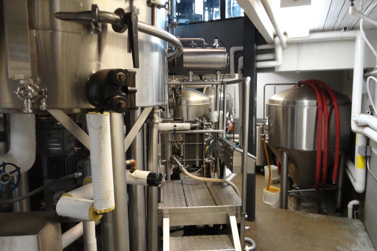 Stainless-steel brewing tanks, pipes, valves, and a short metal platform inside a brewery.