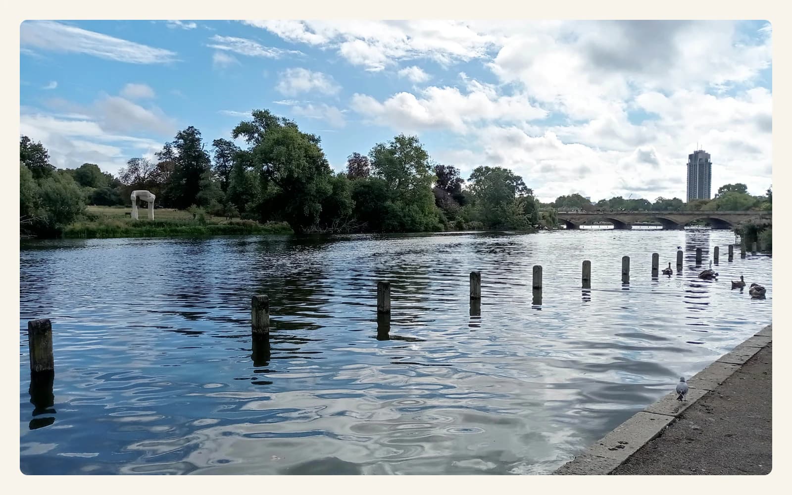The Serpentine in Hyde Park, with water, trees, a low bridge, and a tall building under a partly cloudy sky.