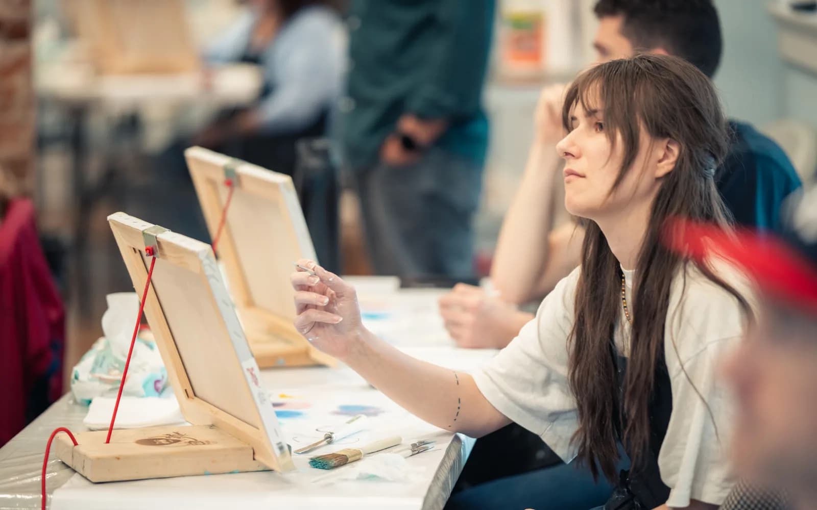 An adult participant paints at a tabletop easel while other workshop participants sit behind her.