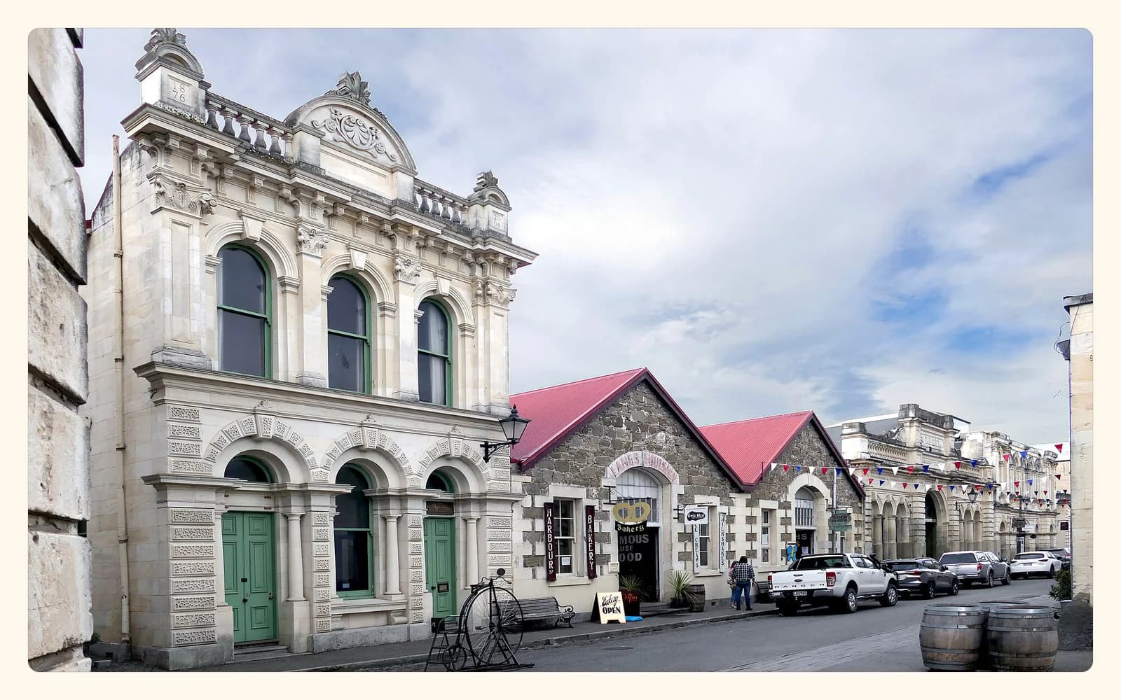 Ornate limestone buildings line Harbour Street in Ōamaru beside a penny-farthing display, shopfronts, and bunting.