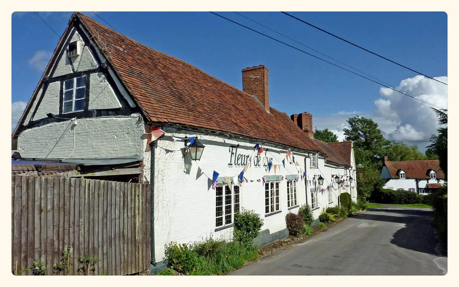 The white-painted Fleur De Lys pub beside a narrow country lane under a blue sky.