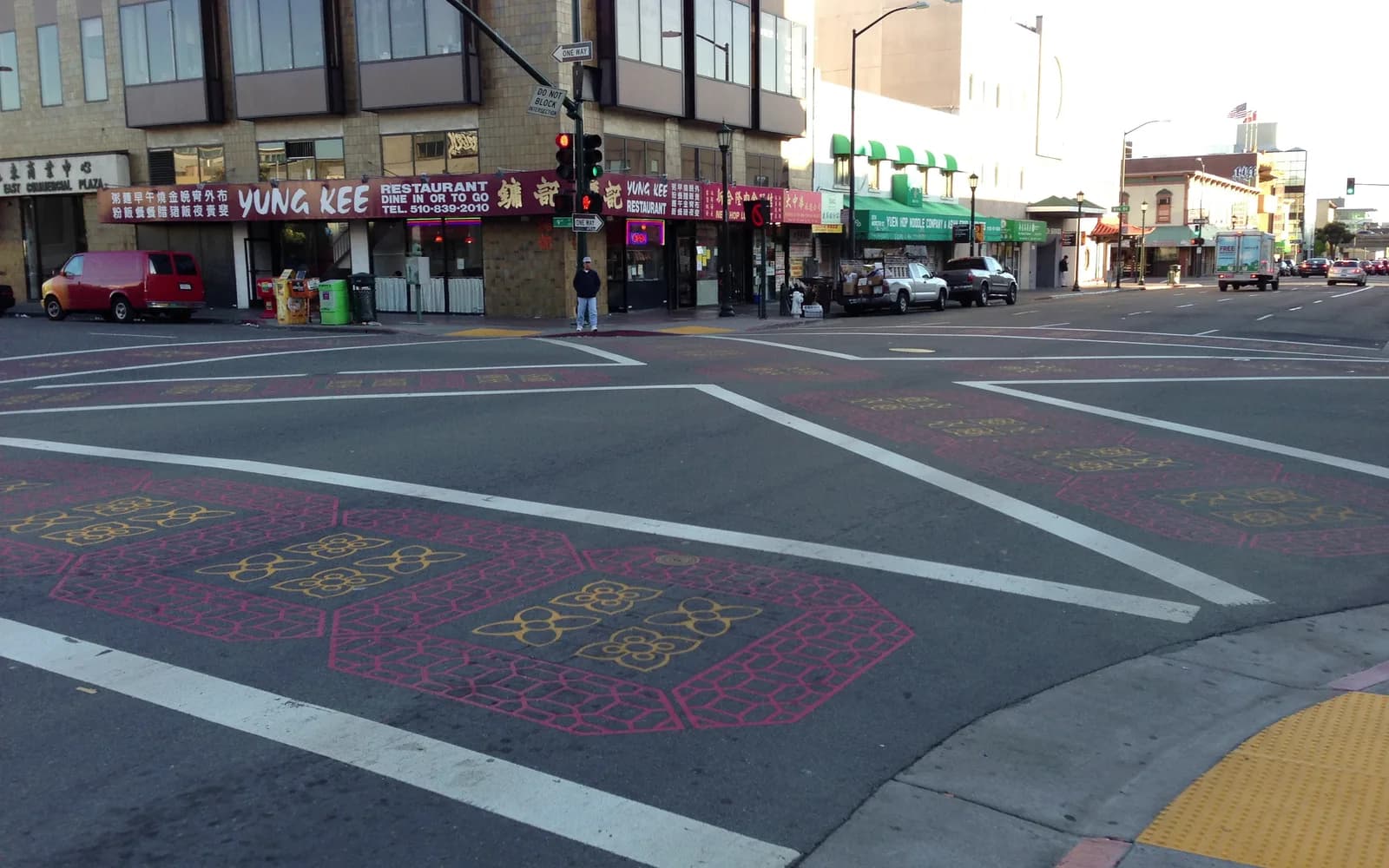 An Oakland street corner with painted crosswalk patterns, shopfronts, and a small number of distant pedestrians.