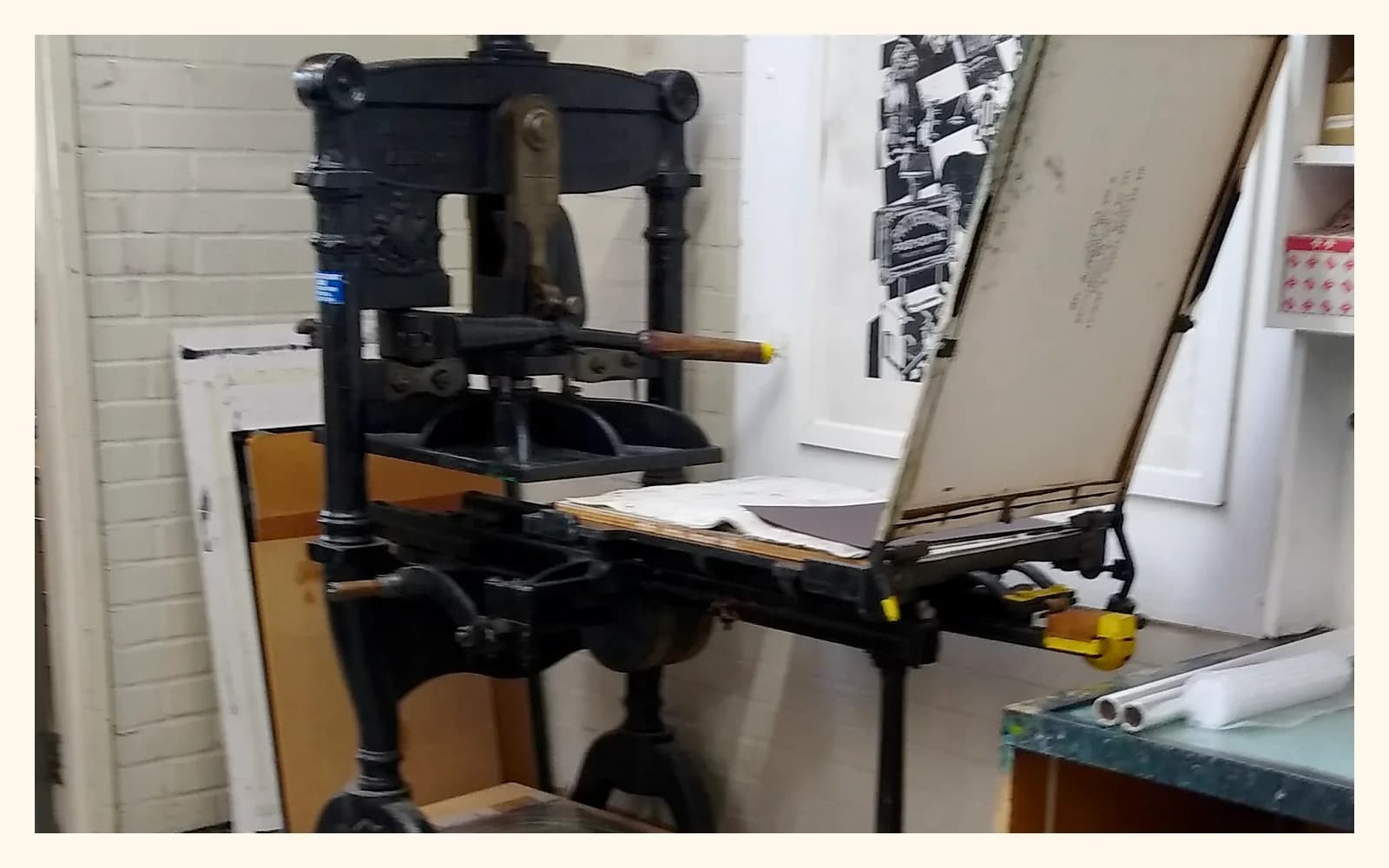 A large black printing press in a bright workshop beside a worktable and framed prints.